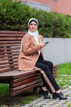 Young woman in hijab uses a smartphone while sitting on a bench outdoors
