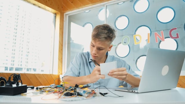 Smart happy boy inspect or learning to use electronic tool while taking a note. Happy highschool student studying about electric equipment and writing comment while sitting at STEM class. Edification.