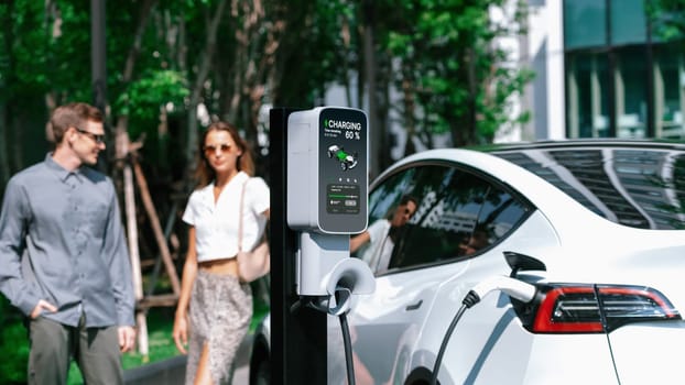 Young couple travel with EV electric car charging in green sustainable city outdoor garden in summer shows urban sustainability lifestyle by green clean rechargeable energy of electric vehicle innards