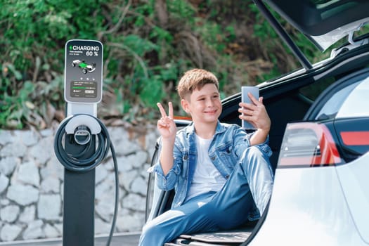 Little boy sitting on car trunk taking selfie using smartphone while recharging eco-friendly car from EV charging station. EV car road trip using sustainable energy concept. Perpetual