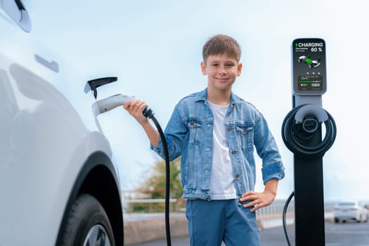 Little boy recharging eco-friendly electric car from EV charging station. EV car road trip travel by the seashore by alternative vehicle powered by clean renewable and sustainable energy. Perpetual