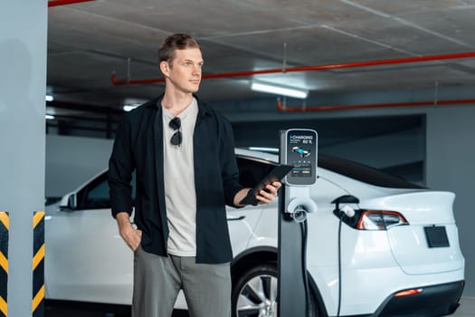 Young man travel with EV electric car to shopping center parking lot charging in downtown city showing urban sustainability lifestyle by green clean rechargeable energy of electric vehicle innards