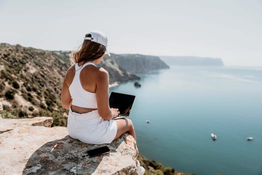 Digital nomad, Business woman working on laptop by the sea. Pretty lady typing on computer by the sea at sunset, makes a business transaction online from a distance. Freelance remote work on vacation