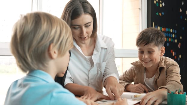 Professional caucasian teacher telling story to diverse student while sitting at table with storybook and colored book. Smart learner listening story while colored picture from instructor. Erudition.