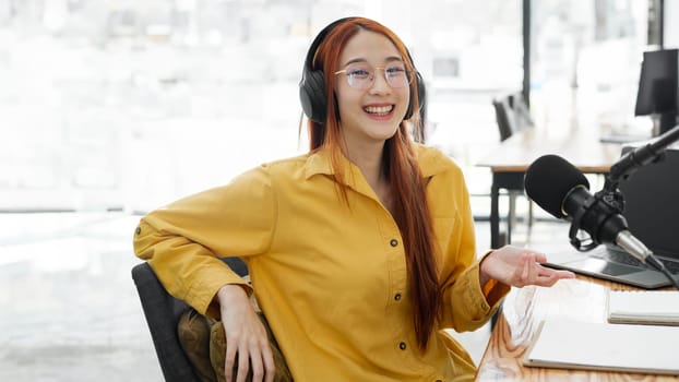 Cheerful woman hosting a live podcast, engaging with audience using professional microphone in studio.