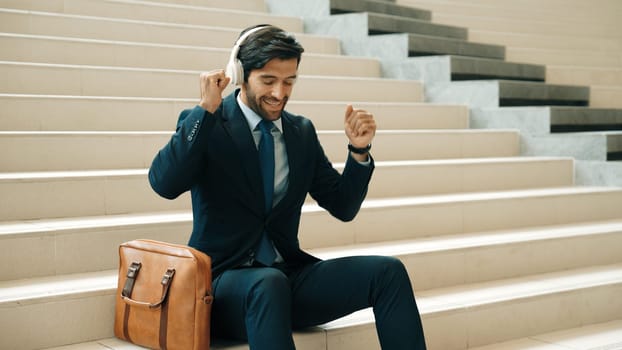 Professional business man picking and listening music by using headphone while sitting at stairs. Project manager dance while listening song while wearing headphone, suit. White background. Exultant.