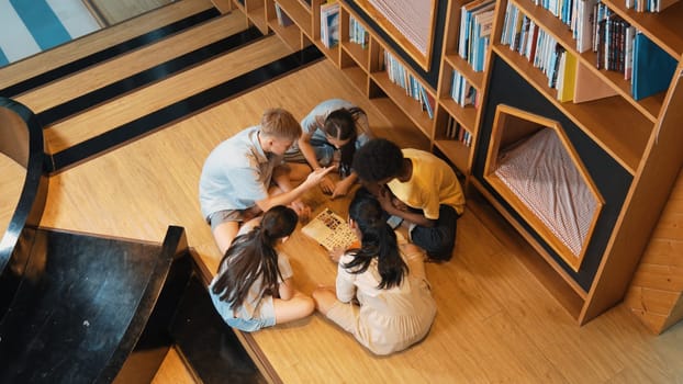 Top view of diverse children sitting in circle while looking at camera and waving hands at library surrounded with the stack of book. Multicultural highschool student greeting camera. Edification.