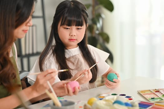 Cute little child girl painting Easter eggs with mother in living room, getting ready for holiday.