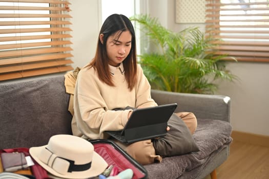 Young Asian woman sitting on couch with suitcase and using laptop for booking hotel online.
