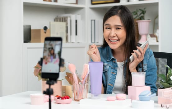 Young beautiful Asian reviewing cosmetic makeup and skin care collection, talking customer on social media online, presenting product surrounded beauty merchandise recording blurred phone . Stratagem.