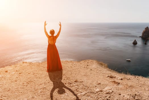 Side view a Young beautiful sensual woman in a red long dress posing on a rock high above the sea during sunrise. Girl on the nature on blue sky background. Fashion photo.