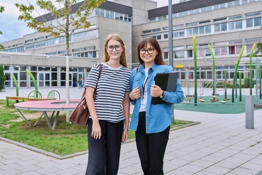 Teacher and teenage schoolgirl looking at camera together outdoor, school building background. Meeting communication student girl with backpack and mentor counselor. Education, adolescence, learning concept