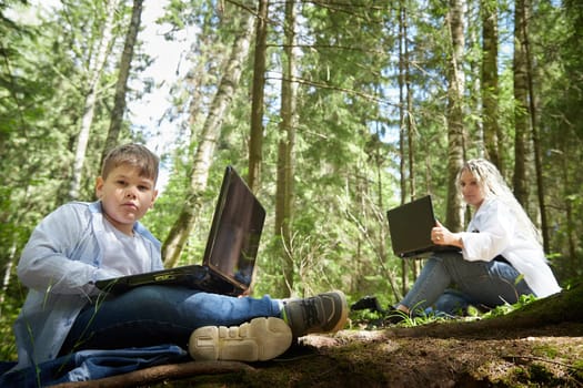 Mother and son with a laptops in the forest in summer. Fat young smart teenage boy and woman working with modern IT technologies in nature