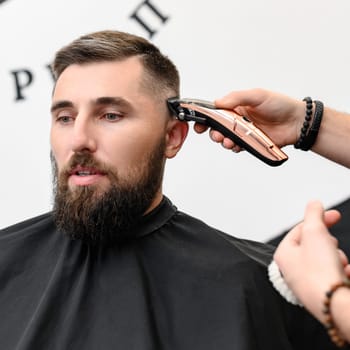 A barber cuts a man with a beard in a barber shop. Short haircut of the client with a clipper.