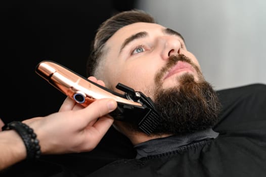 Cutting a gentleman beard in a barbershop with a clipper. Shortening the length of the beard from the sides by the master for the client.