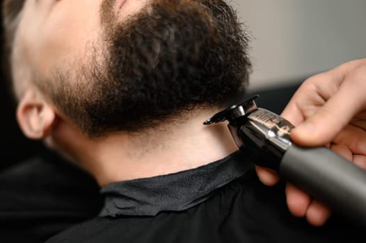 A barber in the salon trims the beard of a Caucasian man with a trimmer in the neck area