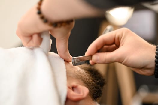 A barber shaves the cheek of a bearded customer with a dangerous razor. Shaving the contour of the beard for the correct shape.