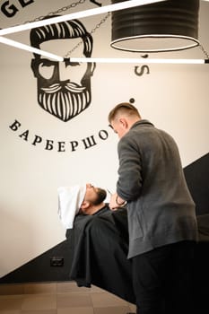 A barber stylist trims the beard of a Caucasian man, whose face is covered with a towel, with scissors