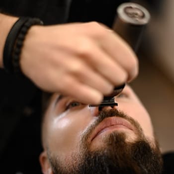 A barber trims a Caucasian client mustache with a trimmer.