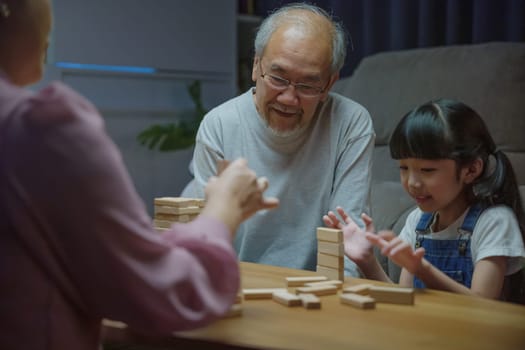 Happy grandparents Asian family enjoy playing toy block with little daughter and mother together at home night time, Smiling parent having fun play build constructor tower of wooden blocks, education