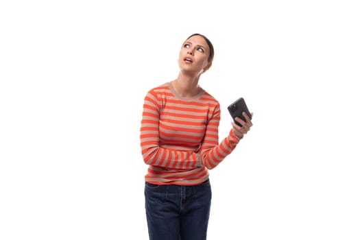 young adult woman dressed in an orange striped sweater looks up dreamily holding a phone.