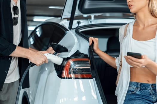 Young couple travel with EV electric car to shopping center parking lot charging in downtown city showing urban sustainability lifestyle by green clean rechargeable energy of electric vehicle innards