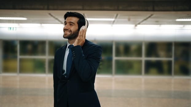 Smart business man listening music by headphone while waiting train at train station with blurring background. Skilled project manager enjoy listen relax sound while holding mobile phone. Exultant.