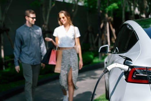 Young couple travel with EV electric car charging in green sustainable city outdoor garden in summer shows urban sustainability lifestyle by green clean rechargeable energy of electric vehicle innards