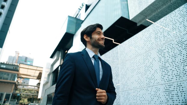 Smart business man wearing suit and smiling while walking at street near white modern architectural building. Attractive handsome manager looking skyscraper in urban city. Copy space. Exultant.