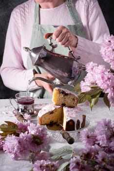 a woman pours tea from a silver teapot against a background of pink sakura flowers, a beautiful Easter cake stands on the table. High quality photo
