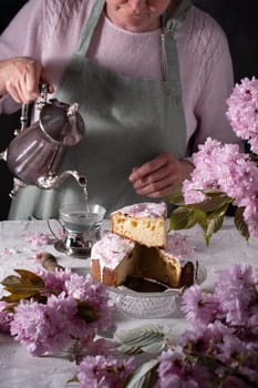a woman pours tea from a silver teapot against a background of pink sakura flowers, a beautiful Easter cake stands on the table. High quality photo