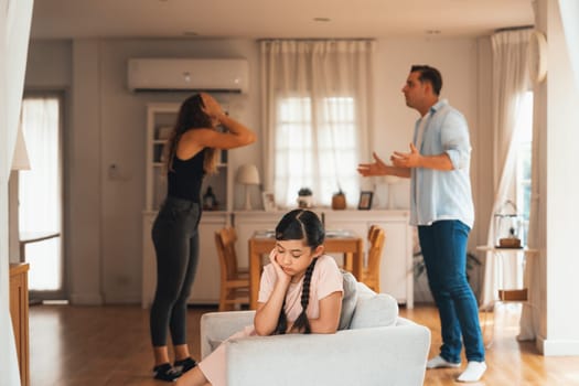 Annoyed and unhappy young girl sitting on sofa trapped in middle of tension by her parent argument in living room. Unhealthy domestic lifestyle and traumatic childhood develop to depression.Synchronos