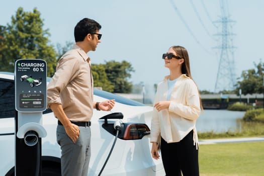 Young couple recharge EV car battery at charging station connected to power grid tower electrical industrial facility as electrical industry for eco friendly vehicle utilization. Expedient