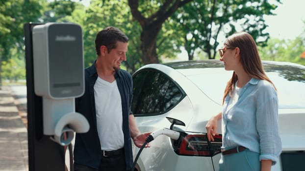 EV car recharging electricity for battery by lovey couple traveling during their road trip travel by eco friendly electric vehicle in national park and greenery forest on holiday. Exalt