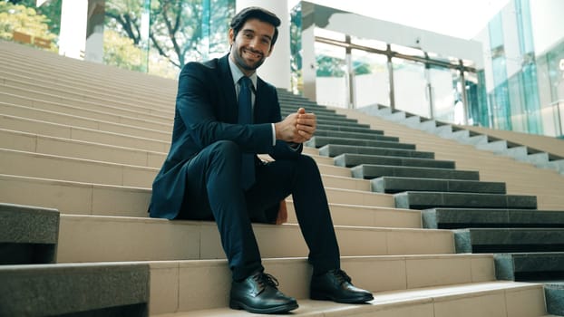 Smiling skilled businessman looking at camera while sitting on stairs. Young professional project manager smile at camera while holding mobile phone at outdoor with blurred background. Exultant.