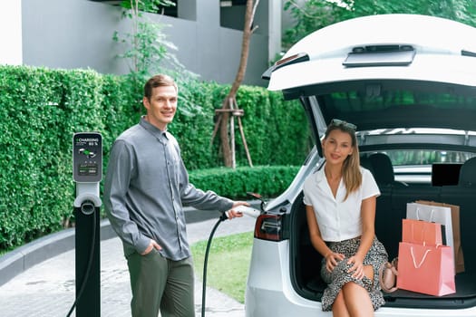 Young couple travel with EV electric car charging in green sustainable city outdoor garden in summer shows urban sustainability lifestyle by green clean rechargeable energy of electric vehicle innards