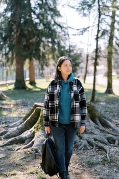 Young lady with a backpack in her hand walks through a coniferous forest and looks away. High quality photo