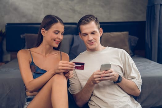 Young couple sit in the home bedroom using online payment app and digital wallet on smartphone to pay with credit card. E commerce shopping and modern purchasing via mobile internet. Unveiling