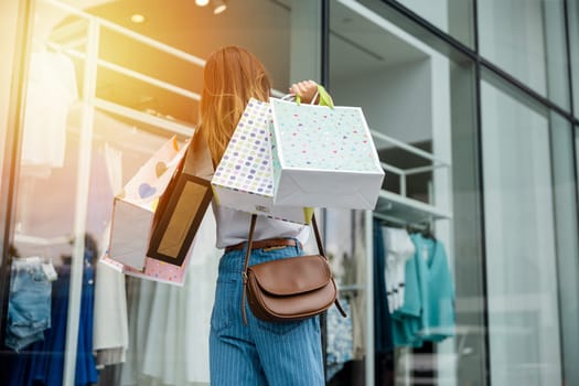 Back view of young woman holding shopping bags and walking in front of a clothing store, enjoying a summer day of retail therapy. She is a trendy consumer, always looking for the latest fashion items.