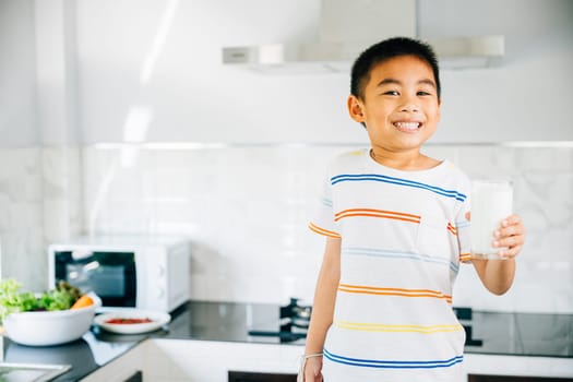 Smiling Asian little boy in kitchen holds milk cup. Portrait of cute son enjoying drink. Joyful child sips calcium-rich liquid, radiating happiness at home, Daily life health care Medicine food