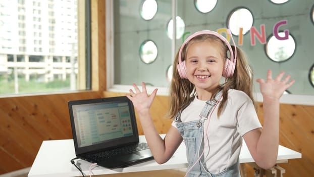 Smiling girl looking while waving hand at camera with laptop placed on table. Child wearing headphone smiling while laptop screen show system programing or coding program in STEM class. Erudition.