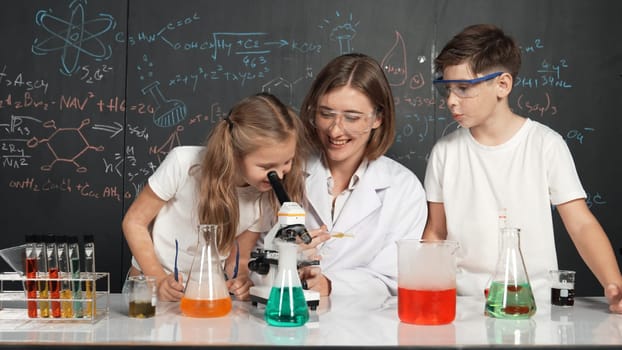 Caucasian boy looking under microscope while teacher giving advice. Professional instructor wearing lab suit looking for diverse student at table with beaker filled with colored solution. Erudition.