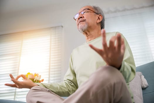 Lifestyle calm senior man lotus pose doing yoga for mental balance breathing air relaxing on sofa at home, Asian elderly old man sitting practise yoga and meditation in lotus position and closed eyes