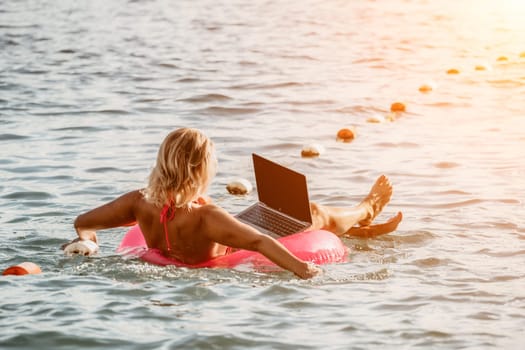 Woman works on laptop in sea. Freelancer, young blond woman in sunglases floating on an inflatable big pink donut with a laptop in the sea at sunset. Freelance, travel and holidays concept