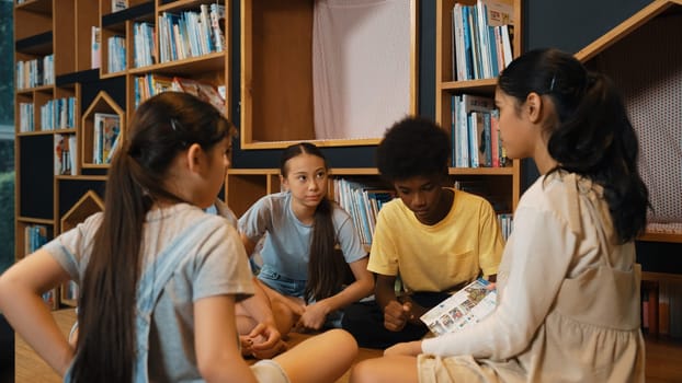 Group of diverse children sitting in circle while reading a book at library. Highschool girl sharing a magazine while pointing at interested topic and talking with multicultural friends. Edification.