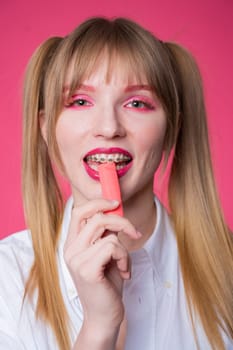 Portrait of a young woman with braces and bright makeup chewing gum on a pink background. Vertical photo