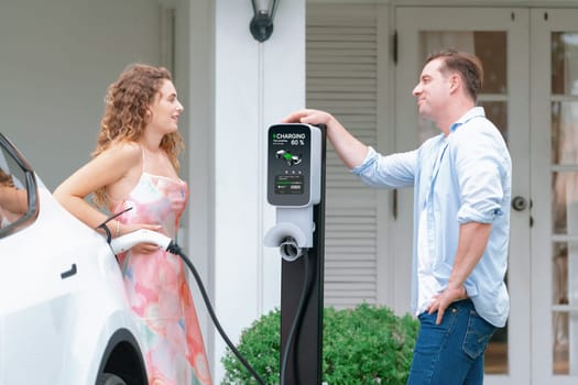 Happy and lovely couple with eco-friendly conscious recharging electric vehicle from EV home charging station. EV car technology utilized for residential home to future sustainability. Synchronos