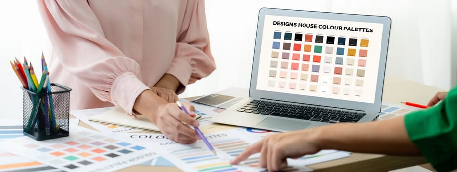 Creative graphic design presents selective color while manager using laptop comparing suitable color at table with color palettes, graphic material scatter around at modern office. Variegated.