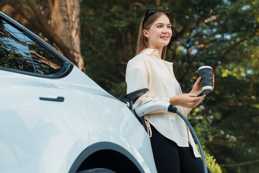 Young woman use smartphone to pay for electricity at public EV car charging station green city park. Modern environmental and sustainable urban lifestyle with EV vehicle. Expedient