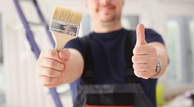 Arms of worker hold paint brush and show confirm sign with thumb up closeup. Manual job DIY inspiration joinery startup idea fix shop hard hat industrial education profession career concept